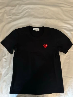 Svart Comme des Garçons PLAY t-shirt - Säljer en svart t-shirt från Comme des Garçons PLAY med det ikoniska röda hjärtat med ögon broderat på bröstet. Klassisk passform, rund hals och korta ärmar. Perfekt basic-plagg med en twist. Tillverkad i mjuk bomull.