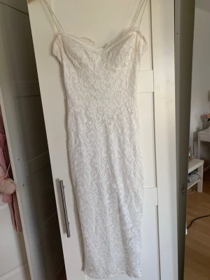 Vit spetsklänning med smala band  - Elegant vit klänning i figurnära modell med vacker spets och off shoulder-skärning. Klänningen har ett romantiskt blommönster i spetsen och en markerad midja. Perfekt för speciella tillfällen där du vill känna dig extra fin. Spetsen på klänningen har släppts lite. 