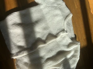 Vit stickad kofta med volang - Supersöt vit stickad kofta med volangdetalj och knappar framtill. Koftan har rund halsringning och ribbade kanter. Perfekt för dig som gillar en mjuk och trendig look.