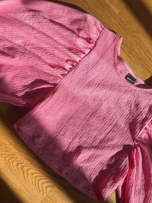 Rosa topp med puffärm från Gina Tricot - Säljer en rosa topp från Gina Tricot med stora puffärmar och strukturerat tyg. Toppen har rund halsringning och en loose passform som ger en cool och trendig look. Perfekt för dig som vill ha en somrig tröja.