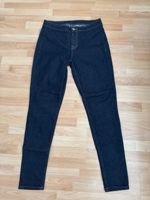 Mörkblå skinny jeans som nya - Snygga mörkblå skinny jeans med hög midja och klassiska fem fickor. Jeansen har kontrastsömmar och en knappstängning framtill. Perfekta för dig som gillar en smal siluett och tidlös stil. Använd en gång. Midjan 76cm.