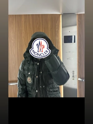 Mörkgrön dunjacka (Maya) från Moncler - Snygg mörkgrön dunjacka från Moncler med glansig finish, hög krage och huva. Jackan har tryckknappar, dragkedja, flera fickor och klassisk Moncler-logga på bröstet. Perfekt för kalla vinterdagar och riktigt varm tack vare dunfyllningen. Priset kan justeras vid smidig affär
