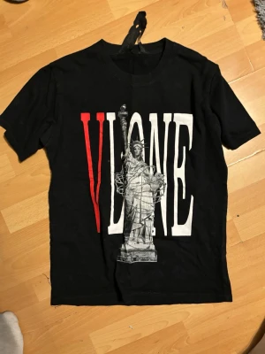 Svart VLONE t-shirt med tryck - Svart t-shirt från VLONE med stort tryck av Frihetsgudinnan och texten 'VLONE' i rött och vitt på framsidan. Baksidan har ett stort rött V och samma statytryck. Perfekt för streetwear-stilen.
