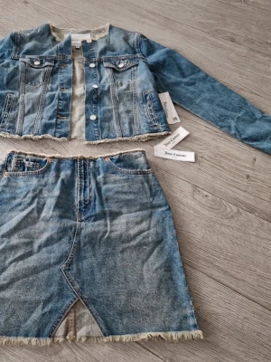 Jeanskjol set med rå kant Juicy Couture M - Säljer en blå jeanskjol från Juicy Couture med råa kanter och fransig nederdel. Kjolen har klassisk femficksdesign, hög midja och en liten slits framtill. Materialet är slitstarkt denim i 100% bomull. Perfekt för en trendig och avslappnad look.