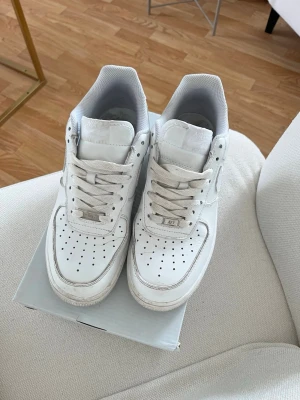 Nike Air Force 1 vita sneakers - Klassiska vita Nike Air Force 1 sneakers i lågt utförande. Skorna har en clean design med perforerad tå, vita skosnören och den ikoniska Swoosh-loggan på sidan. Tillverkade i läder med en stabil gummisula och AIR-detalj på sidan. Perfekta för en stilren och sportig look.