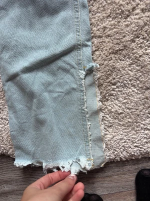 Ljusblå raka jeans med råa kanter - Säljer ett par ljusblå jeans med rak passform och råa, fransiga kanter nertill. Jeansen har hög midja och klassisk femficksdesign. Perfekta för en avslappnad och trendig look. Materialet är denim i bomull.