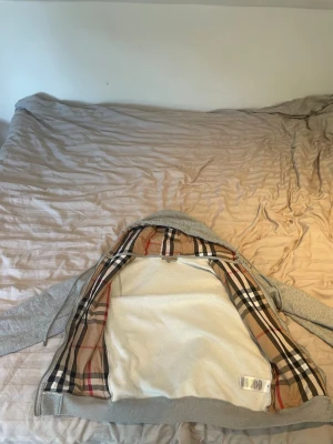 Grå hoodie från Burberry med rutigt foder - Snygg grå hoodie från Burberry med det klassiska beige, röda och svarta rutiga fodret i huvan och insidan. Dragkedja framtill, två fickor och ribbade muddar. Perfekt för dig som vill ha en stilren hoodie med exklusiv känsla och ikoniskt mönster.