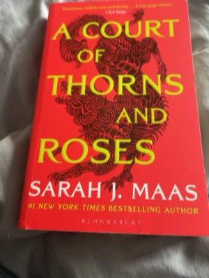A Court of Thorns and Roses - En fantasyroman av Sarah J. Maas med röd och gul omslagsdesign och drakmotiv. Boken är på engelska och handlar om Feyre som dras in i en magisk och farlig värld. Perfekt för dig som gillar romantik, spänning och äventyr.