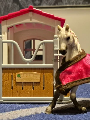 Schleich Leksaksstall med häst och figur - Säljer ett detaljerat leksaksstall i plast med rött tak och träfärgade dörrar. Medföljer en vit häst med rosa täcke och en liten mänsklig figur.