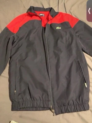  Lacoste Sport track suit - Snygg track suit  från Lacoste Sport i svart med röda axelpartier. Jackan har dragkedja framtill, två sidofickor med dragkedja och elastisk nederkant. Klassisk Lacoste-logga på bröstet och 'LACOSTE SPORT' tryckt på ryggen. Perfekt för en sportig och trendig look.