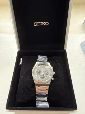 Seiko Cronograph  - Stilren Seiko herrklocka i silver med länkarmband i metall. Urtavlan är silverfärgad med diskreta ränder och tre mindre svarta subdials. Klockan har datumvisning och tydliga indexmarkeringar. Perfekt accessoar för dig som gillar klassisk design. Aldrig använd och kvar i orginalplast!