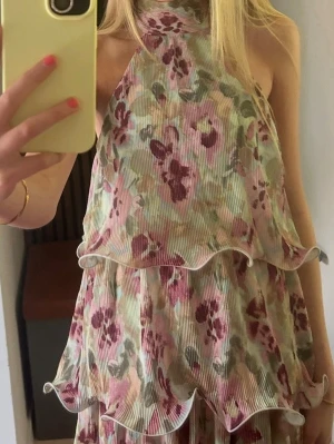 Blommig volangklänning med halterneck - Säljer en söt blommig klänning med volanger och halterneck. Klänningen har ett ribbat tyg i ljusa pastellfärger med rosa och grönt. Använd 1 gång 