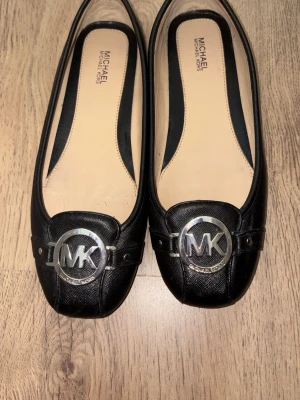 Svarta ballerinas från Michael Kors - Snygga svarta ballerinaskor från Michael Kors i läder med rundad tå och silverfärgad MK-logga framtill. Klassisk design med beige innersula för extra komfort. Perfekta för dig som gillar stilrena detaljer. Har används 1 gång 
