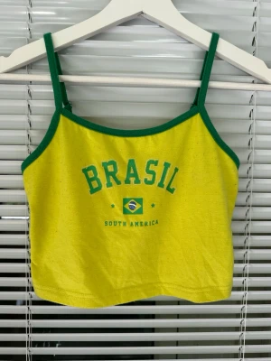 Gul Brasil linnetopp med gröna detaljer storlek: S - Cool gul linnetopp med gröna kanter och smala axelband. Tryck med texten 'BRASIL' och en brasiliansk flagga på framsidan. Perfekt för dig som vill sticka ut och visa din kärlek till Brasilien. Materialet känns som bomull och toppen är croppad. Linnet är nopprig.
