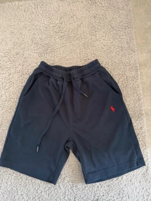 Mörkblå shorts från Polo Ralph Lauren - Snygga mörkblå shorts från Polo Ralph Lauren med elastisk midja och snörning. Klassisk röd logga broderad på vänster ben. Mjukt material och två sidofickor gör dem perfekta för chill dagar. Passar till allt och är riktigt bekväma.