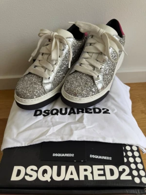 Glittriga sneakers från Dsquared2 - Snygga sneakers från Dsquared2 täckta i silverglitter med vita detaljer och breda vita skosnören. Skorna har en vit sula med svart kant och en rosa detalj på hälen. Insidan är svart och tungan är vit med Dsquared2-logga. Perfekta för dig som vill sticka ut. Använd 4 gånger sparsamt, box och dustbag medföljer 