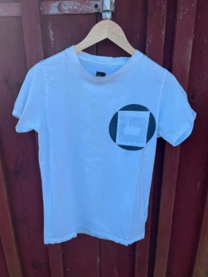 Albam Vit T-Shirt med Bröstficka – Strl S - Vit t-shirt från Albam i bomull med svart cirkeltryck och bröstficka på framsidan. Klassisk rund hals och korta ärmar. Perfekt för en avslappnad och stilren look.