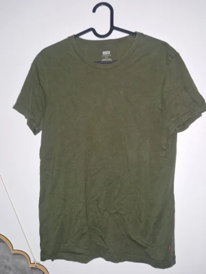 Tshirt Levis - Säljer en enkel och stilren olivgrön t-shirt från Levi's. T-shirten har rund halsringning, korta ärmar och en liten röd Levi's-tag på sidan. Tillverkad i mjuk bomull som känns skön mot huden. Perfekt basplagg för alla tillfällen. Inga defekter
