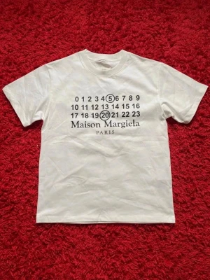 Maison Margiela vit t-shirt - Vit t-shirt från Maison Margiela med det ikoniska siffertrycket och logotypen på bröstet. Klassisk rund hals och korta ärmar. Tillverkad i mjuk bomull för en skön och avslappnad look.