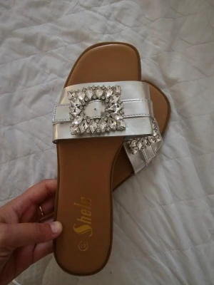 Silverfärgade sandaler med strass - Säljer ett par snygga sandaler från Shelbi i silverfärgat material med bred rem och stora, glittrande strassdetaljer framtill. Sandalerna har en fyrkantig tå och platt sula i brunt. Perfekta för att ge din outfit lite extra bling.