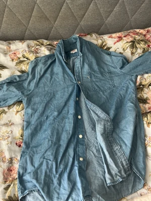 Ljusblå jeansskjorta med bröstficka - Säljer en ljusblå jeansskjorta i bomull med klassisk krage, knappar framtill och en bröstficka. Skjortan har lång ärm och avslappnad passform, perfekt för en casual look. Passar dig som gillar denim och enkel stil.