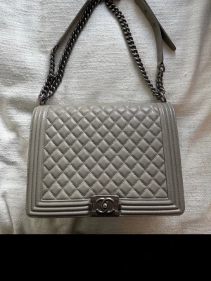 Grå väska i läder - Säljer en grå Boy Bag i quiltat läder med silverfärgad kedjerem och ikoniskt spänne framtill. Väskan har en boxig form och exklusiv känsla, perfekt för dig som vill ha en klassisk men edgy accessoar.