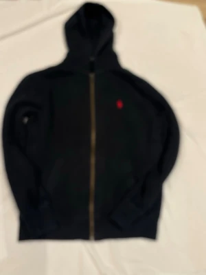 Svart hoodie med dragkedja från Polo Ralph Lauren - Säljer en svart hoodie med dragkedja från Polo Ralph Lauren. Klassisk design med huva och fickor framtill. Liten röd logga broderad på bröstet. Perfekt för en avslappnad och stilren look.