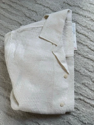 Vit kortärmad skjorta i linne-look - Säljer en stilren vit skjorta med korta ärmar och klassisk krage. Skjortan har ett diskret strukturerat mönster i tyget och knäpps med ljusa knappar framtill. Perfekt för en clean och fräsch sommarstil. Nypris 429kr