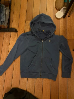 Blå hoodie från Polo Ralph Lauren - Snygg blå hoodie från Polo Ralph Lauren med dragkedja framtill och klassisk huva. Hoodien har två fickor på magen och den ikoniska loggan broderad på bröstet. Tillverkad i mjukt bomullsmaterial som är skönt att ha på sig. Priset kan diskuteras. Nypris 2200. Knappast använd.