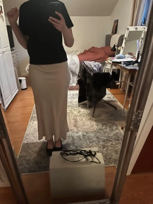 Beige långkjol i satin - Säljer en stilren beige långkjol i satinliknande material. Kjolen har en mjuk, lätt glansig yta och faller snyggt längs kroppen. Perfekt för dig som gillar minimalistisk och elegant stil.