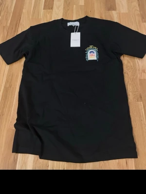 Svart Casablanca t-shirt med tryck - Svart t-shirt från Casablanca med färgglatt tryck på bröstet och stort grafiskt motiv på ryggen. Klassisk passform och rund halsringning. Tillverkad i mjuk bomull, perfekt för en avslappnad och trendig stil.