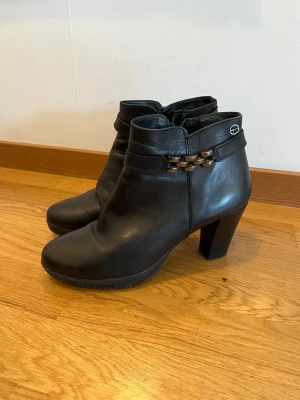Svarta stövletter med klack och kedjedetalj - Snygga och bekväma ankelboots från Tamaris i svart läder. Modellen har en stabil klack, dragkedja på sidan och dekorativ kedjedetalj som ger skorna en elegant och tidlös look.  Perfekta till höst, vinter och vår och passar lika bra till jeans som klänning eller kostymbyxor.