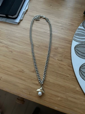 Marianilsdotterhalsband bumble bee - Snyggt halsband med grov silverfärgad kedja och ett hänge i form av en guldfärgad bi med en vit pärla som kropp. Perfekt för dig som gillar unika detaljer och vill sticka ut lite extra. Passar till många olika stilar och är lätt att matcha med andra smycken.