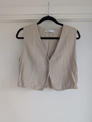 Croppad linneväst x Zara - Stilren beige väst från Zara i linne med v-ringad hals och knäppning framtill. Västen har en croppad passform och är perfekt för att skapa en trendig lager-på-lager-look. Enkel och clean design som passar till många outfits.