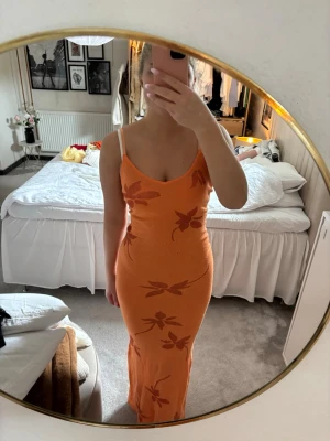 Orange långklänning med blommor - Trendig orange långklänning med smala axelband och blommigt mönster i en mörkare orange ton. Klänningen har en figurnära passform och är perfekt för sommarens alla tillfällen. Materialet känns mjukt och stretchigt, troligen bomull eller syntet. Klänningen är i bra skick då den inte är använd många gånger🧡