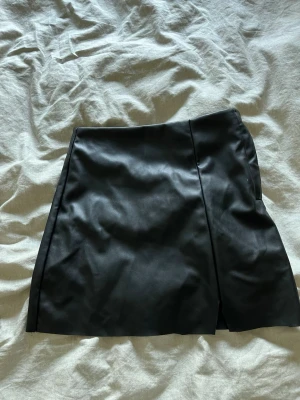 Svart kortkjol i skinnimitation - Snygg svart kortkjol i skinnimitation med en stilren design och diskret slits framtill. Kjolen har en lätt A-linjeformad siluett och stängs med dragkedja i sidan. Perfekt att matcha med en cool t-shirt eller stickad tröja.