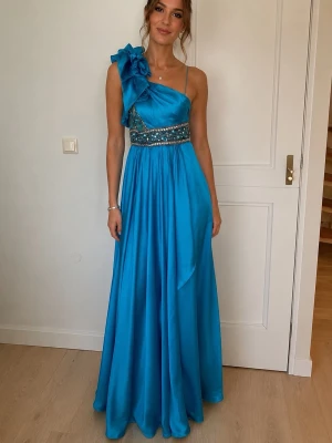Blå turkos långklänning med rosett - Säljer en blå turkos  långklänning i glansigt material med en stor rosett på ena axeln och dekorativa pärlor i midjan. Klänningen har oneshoulder-design och ett elegant fall. Perfekt för bal eller festliga tillfällen.