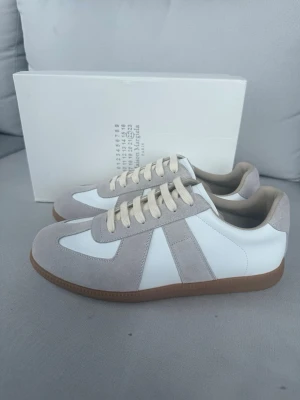 Maison Margiela  sneakers - Maison Margiela  sneakers i vitt och ljusgrått med ovandel i läder och mocka. Klassisk låg modell med beige snörning och brun gummisula. Perfekta för dig som gillar minimalistisk och clean stil.