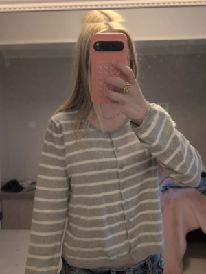 Gråvit randig stickad cardigan - Mysig stickad cardigan i grått och vitt med horisontella ränder. Tröjan har knappar framtill, rund halsringning och långa ärmar. Perfekt att slänga över en t-shirt för en avslappnad look. Materialet känns mjukt och lätt.