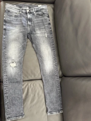 Mörk grå jeans från Calvin Klein - Säljer ett par grå tvättade jeans från Calvin Klein Jeans med skinny fit och slitningar på benen. Jeansen har klassisk femficksmodell, dragkedjegylf och tydliga slitdetaljer för en trendig look. Materialet är bomull med stretch för skön passform.