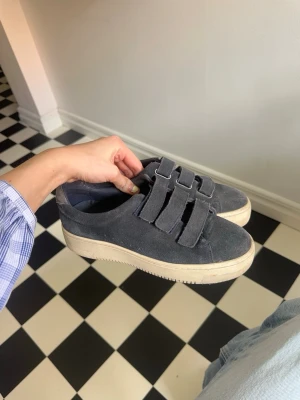 Sandro mocka sneakers med kardborre - Marinblå sneakers från Sandro Paris med kardborreband i stilren design. Skorna är i bra skick men har synliga tecken på användning, och har inte använts på flera år. Originalkartong medföljer.