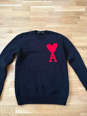 Svart stickad tröja Ami Paris - Svart stickad tröja från Ami Paris med röd hjärtlogga och bokstaven A på bröstet. Tröjan har rund halsringning, långa ärmar och ribbade muddar vid ärmslut och nederkant. Perfekt för dig som gillar stilrena plagg med en twist. Den har legat i garderoben ett tag och vill bli av med den 