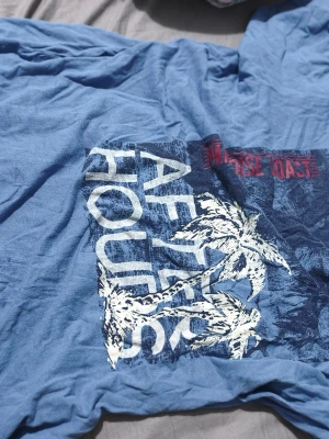 Blå t-shirt med palmtryck från Livergy - Säljer en blå t-shirt från Livergy med stort tryck på framsidan som visar palmer och texten 'AFTER HOURS'. T-shirten har korta ärmar och är gjord i mjuk bomull, perfekt för en avslappnad stil. 3 tröjor