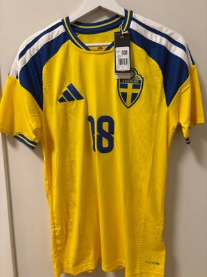 Sverige matchtröja Ayari #18 Adidas - Gul och blå officiell Sverige matchtröja från Adidas med nummer 18 och namnet Ayari på ryggen. Tröjan har blå detaljer på axlar och ärmar, samt Sveriges landslagsemblem på bröstet. Tillverkad i lätt och ventilerande funktionsmaterial. Helt oanvänd och du får den i orginalbox. Beställde x2, därför säljer jag nu den ena. Ny pris:1400kr.