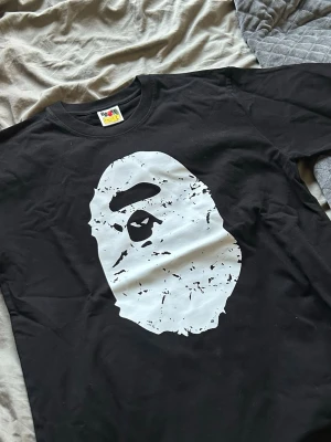 Svart BAPE t-shirt med stor logga - Säljer en svart t-shirt från BAPE med den ikoniska vita apansiluetten tryckt på framsidan och stor 'BATHING APE' text på ryggen. Klassisk rund hals och korta ärmar. Perfekt för dig som gillar streetwear och vill sticka ut.