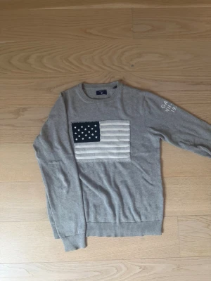 Grå stickad tröja med USA-flagga - Snygg grå stickad tröja från Gant med amerikansk flagga i vitt och blått på bröstet. Rund halsringning och ribbade muddar vid ärmslut och nederkant. Diskret Gant-brodyr på ena ärmen. Perfekt för en avslappnad men stilren look.