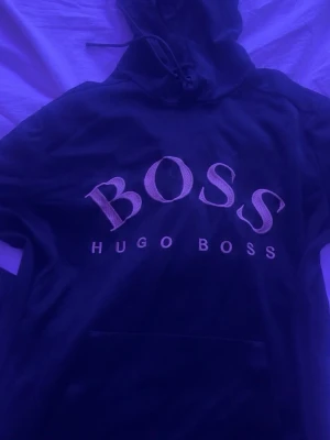 Svart hoodie från Hugo Boss - Snygg svart hoodie från Hugo Boss med stor BOSS-logga broderad på bröstet och klassisk känguruficka. Huva med dragsko och mjukt material som känns skönt mot huden. Perfekt för en avslappnad och trendig stil.