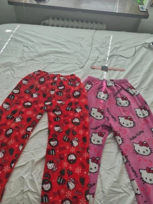 Hello Kitty fleecepyjamasbyxor - Två par supermjuka fleecepyjamasbyxor från Hello Kitty. Ena paret är rött med juliga Hello Kitty-motiv, snöflingor och rosetter, det andra är rosa med stora Hello Kitty-ansikten och text. Perfekta för mysiga hemmakvällar och vinterchill.