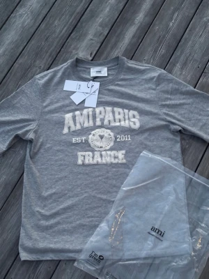 Grå t-shirt från Ami Paris - Snygg grå t-shirt från Ami Paris med vit text och emblem på bröstet. Klassisk rund halsringning och korta ärmar. Materialet är mjuk bomull och designen har en cool collegekänsla med tryck 'AMI PARIS FRANCE EST 2011'. Perfekt för dig som gillar stilrena plagg.