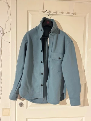 Ljusblå overshirt från H&M - Snygg ljusblå overshirt från H&M med klassisk krage, tryckknappar och en stor bröstficka med lock. Overshirten har raka ärmar och är tillverkad i mjukt material som passar perfekt för lager på lager-stil.